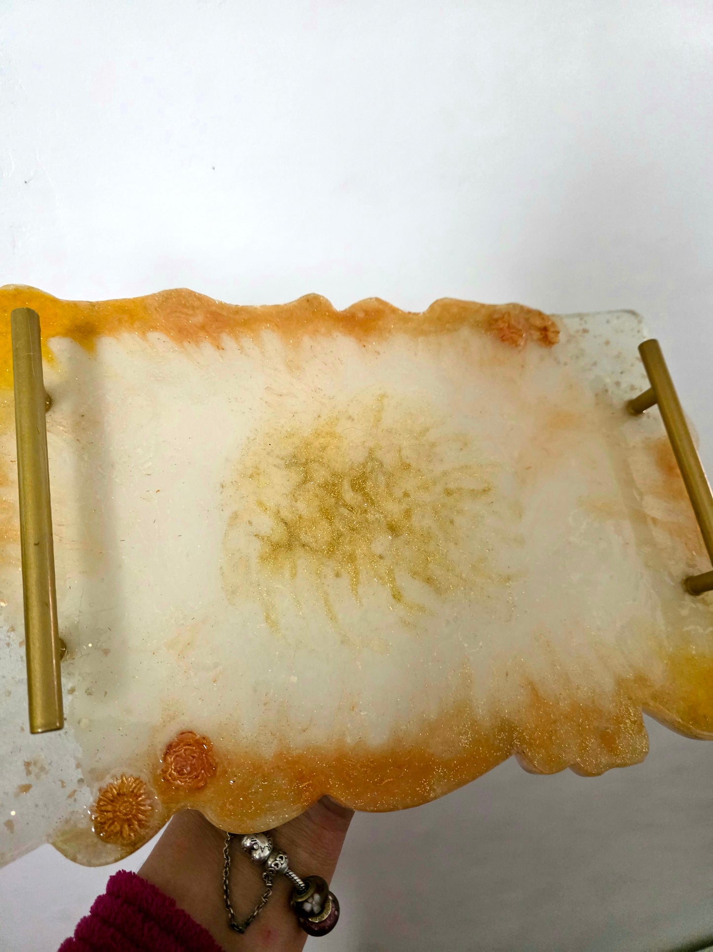 A rectangular resin tray with a wavy, organic border, featuring a gold metal handle on top and a gold metal accent on the bottom right. The tray is held by a hand, showing a hand-painted design with a white center, golden-brown sunburst patterns, and orange accents.