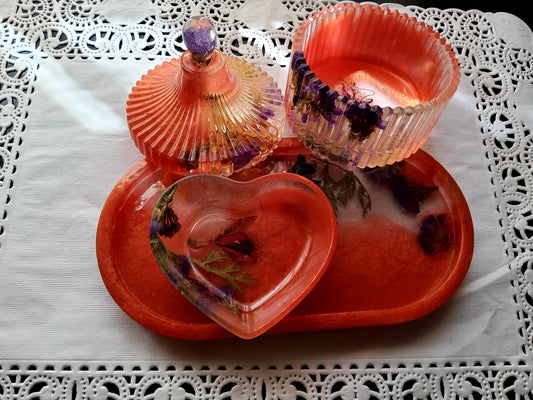 A set of decorative resin keepsake items in a warm coral-orange hue with embedded purple and green flowers, arranged on a matching oval tray over a white lace doily.